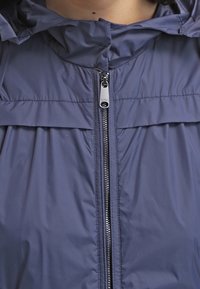 Coupe-vent bleu marine avec fermeture éclair à l'avant, capuche et détail plissé aux épaules. Fabriqué en matière synthétique légère et lisse.