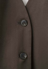 Tissu marron texturé avec deux boutons sombres, ronds et à quatre trous, fixés sur le devant d’un blazer ou d’un manteau.