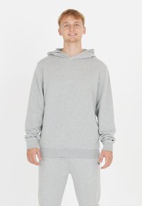 Ung man med ljust hår som ler, iklädd en ljusgrå huvtröja och matchande träningsbyxor, stående mot en vit bakgrund.