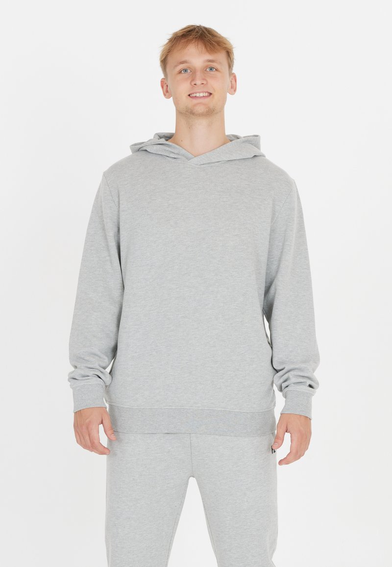 Ung man med ljust hår som ler, iklädd en ljusgrå huvtröja och matchande träningsbyxor, stående mot en vit bakgrund.