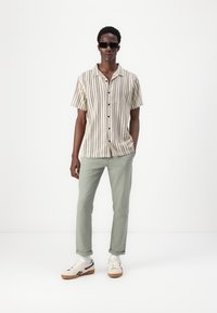 Short-sleeve striped shirt in beige and cream, paired with light green pants. Model wears dark sunglasses and beige sneakers.