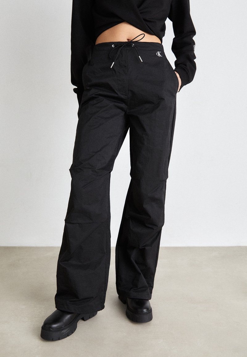 Personne portant un pantalon noir à jambes larges avec une taille à cordon et des bottes noires épaisses, debout sur un sol gris clair contre un mur blanc.