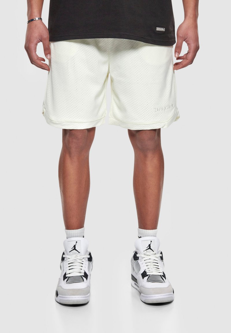 Shorts de sport en maille de couleur jaune clair, avec une surface texturée et un logo brodé, accompagnés de baskets blanches et noires.