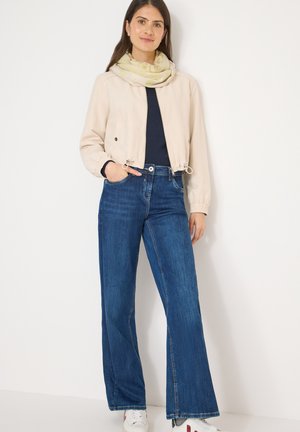 Veste beige courte avec taille à cordon sur une chemise marine, associée à un jean large bleu et des baskets blanches, agrémentée d'une écharpe légère à motifs.