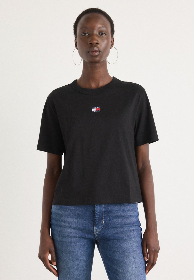 Tricou negru din bumbac, cu mâneci scurte și guler rotund. Prezintă un mic logo în roșu �și albastru. Este purtat cu blugi albaștri.