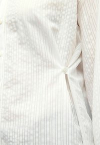Chemise blanche à boutons avec de subtiles rayures verticales, tissu texturé et détail noué sur le côté à la taille, dotée de fermetures à boutons classiques.