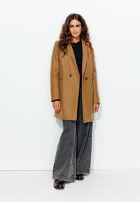Manteau en laine marron au design ajusté, avec deux boutons, porté sur un haut noir, associé à un jean large taille haute gris et des chaussures marron.