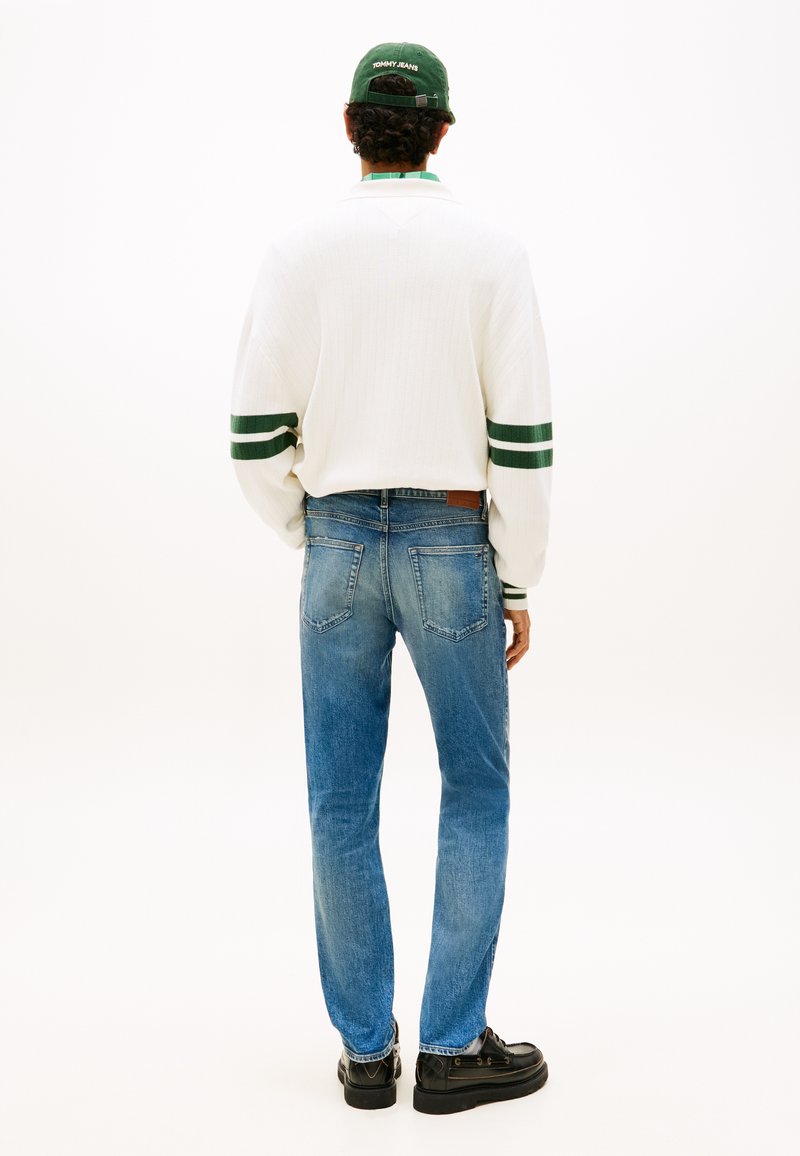 Homme portant un jean bleu, un pull blanc à rayures vertes, une casquette verte et des chaussures noires, debout, dos à la caméra.