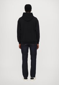 Persona con una sudadera negra con diseños de llamas en las mangas, jeans oscuros y zapatillas blancas, de pie frente a un fondo blanco liso.