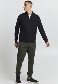 Pulover negru cu nervuri, cu guler cu fermoar, asortat cu pantaloni verzi măsliniu, conici și adidași negri, având o croială casual și o textură tricotată.