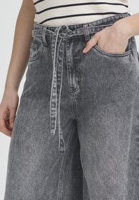 Personne portant un jean noir délavé taille haute avec une ceinture en tissu nouée en nœud et une main dans la poche avant.