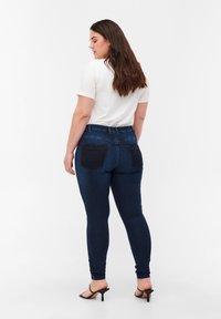 Jeans oscuros ajustados con una cintura de media altura, bolsillos traseros con parche y un leve estiramiento, combinados con una camiseta blanca y sandalias negras de tacón.