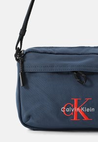 Bolso bandolera de nailon azul con herrajes negros, cierre de cremallera y texto de logo bordado en rojo "Calvin Klein" y "CK". Superficie texturizada.