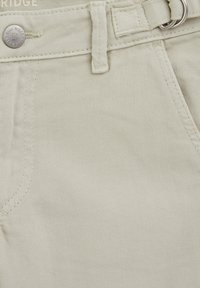 Nærbillede af beige denim bukser, der viser frontlomme, bæltestrop, knap med "Blue Ridge" indgravering og dobbeltd-rings bælte detalje.