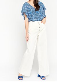 Blusa blu a motivi con maniche a volant, abbinata a pantaloni bianchi a gamba larga con chiusura a cinque bottoni e sandali blu.