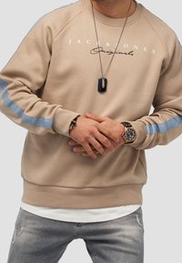 Beige sweatshirt med blå ränder på ärmarna, rundad halsringning och logotext. Matchad med grå slitna jeans och accessoarer, inklusive en klocka.