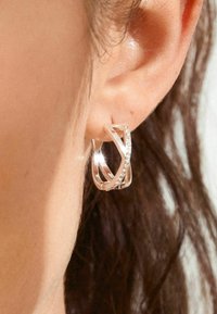 Pendientes de aro en oro rosa con formas entrelazadas, que cuentan con pequeñas piedras transparentes a lo largo de un borde, reflejando la luz, llevados en la oreja.