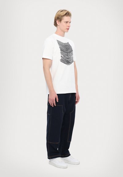 Baltas kokvilnas t-krekls ar melnu lineāru grafisko dizainu, apvienots ar brīviem tumši ziliem biksēm ar izteiksmīgu šuvju un kabatu dizainu, valkājot baltas sporta kurpes.