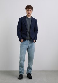 Blazer azul marino sobre un suéter gris, combinado con jeans azul claro y zapatos negros. El blazer tiene dos botones y un corte entallado.