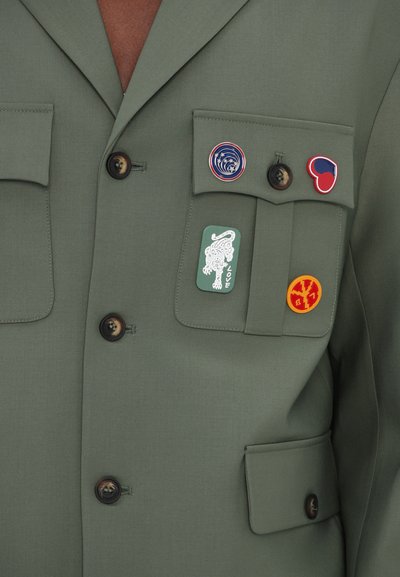 Blazer vert olive avec une finition texturée, comportant deux poches poitrine, quatre boutons et quatre badges colorés avec différents motifs.