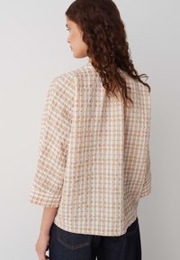 Blouse légère à carreaux beige et blanc avec un motif texturé. Présente une coupe décontractée et des manches trois-quarts.