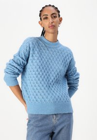 Helles blaues Strickpullover mit strukturiertem Diamantmuster, geripptem Rundhalsausschnitt und Bündchen. Kombiniert mit Jeans, die eine lockere Passform zeigen.
