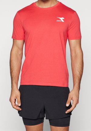 Mężczyzna ubrany w czerwoną sportową koszulkę z krótkim rękawem z białym logo na piersi oraz czarne, warstwowe spodenki, stojący na jednolitym tle.