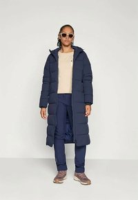 Marinblå vadderad lång kappa med huva, med dragkedja och sidofickor, bärs över en beige topp och marinblå byxor, kompletterad med solglasögon.