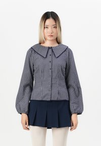 Blusa a scacchi blu navy e bianca con un ampio colletto, chiusura frontale con bottoni e maniche a sbuffo, abbinata a una gonna plissettata blu navy.