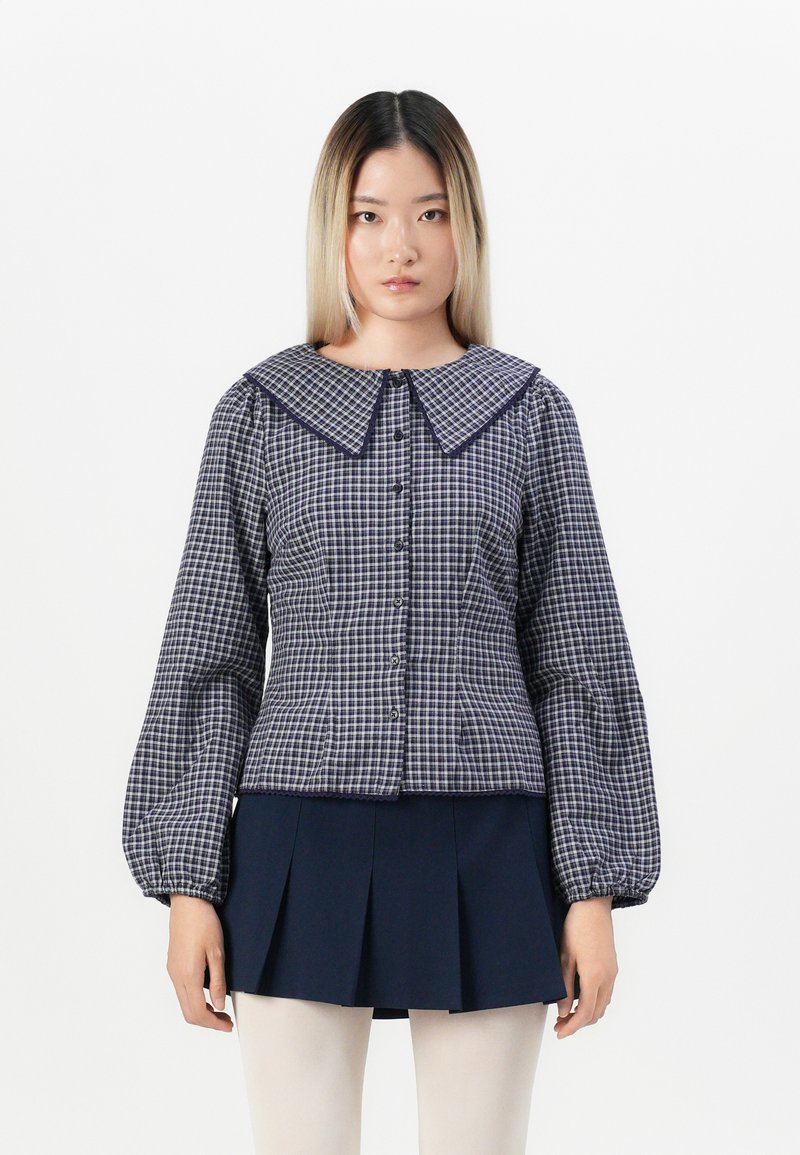 Blusa a scacchi blu navy e bianca con un ampio colletto, chiusura frontale con bottoni e maniche a sbuffo, abbinata a una gonna plissettata blu navy.