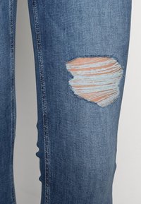 Jeans de mezclilla azules con una zona desgastada y bordes deshilachados en el muslo derecho, mostrando variaciones de color azul claro y oscuro.