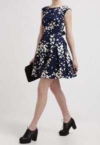 Navy mekko, jossa on valkoinen perhoskuvio, istuva bodice, leveä helma, hihattomat hihat, yhdistettynä mustaan kukkaroon ja laattakenkiin.
