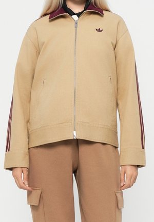 Personne portant une veste beige zippée avec des bandes bordeaux sur les manches et un pantalon cargo marron assorti, debout devant un fond uni.