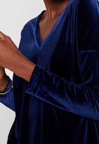 Top in velluto blu navy con design a V, maniche lunghe e una texture soffice, caratterizzato da una vestibilità ampia e senza decorazioni visibili.