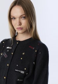 Jonge vrouw met steil blond haar, gekleed in een donkere cardigan versierd met kleine patches, sterren en gouden knopen op een lichte achtergrond.