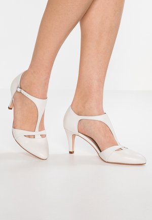 Chaussures à talons hauts en cuir blanc avec un design découpé, un bout pointu et une bride de cheville ajustable. Elles présentent un talon fin et un éclat discret.