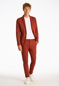 Abito color ruggine realizzato in tessuto liscio con taglio sartoriale. Comprende un blazer con bottone singolo, pantaloni slim-fit e sneakers bianche.