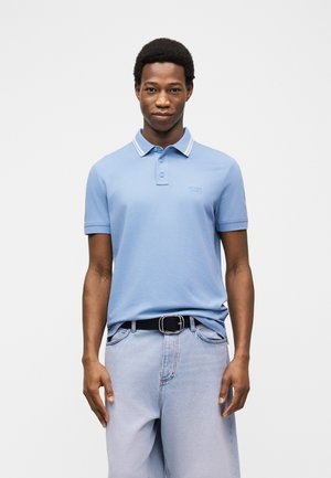 Άνδρας που φοράει ανοιχτό μπλε μπλουζάκι πόλο με κοντά μανίκια και ανοιχτό μπλε τζιν με μαύρη ζώνη, στέκεται μπροστά σε απλό λευκό φόντο.