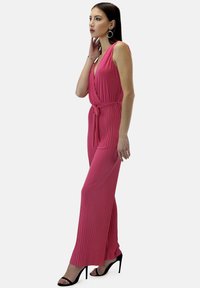 Monosuit plisado de color fucsia con diseño envolvente y tirante a la cintura. Presenta piernas anchas y un escote pronunciado, combinado con tacones altos negros.
