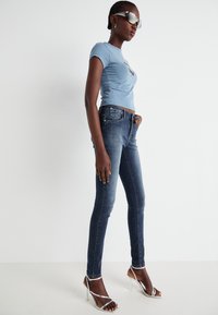 T-shirt aderente azzurro chiaro, jeans skinny blu scuro e sandali alti argentati. Modella con occhiali da sole riflettenti, in piedi contro uno sfondo bianco.