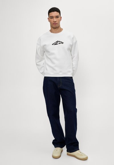 Sweatshirt blanc avec un logo noir, associé à un jean bleu foncé et des baskets beiges. Le sweatshirt présente un col rond et des poignets côtelés.