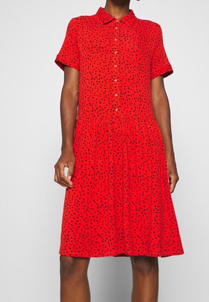 Kobieta ubrana w czerwoną sukienkę na krótki rękaw z guziczkami, z czarnym nieregularnym wzorem w kropki i kołnierzykiem, stojąca na jednolitym tle.