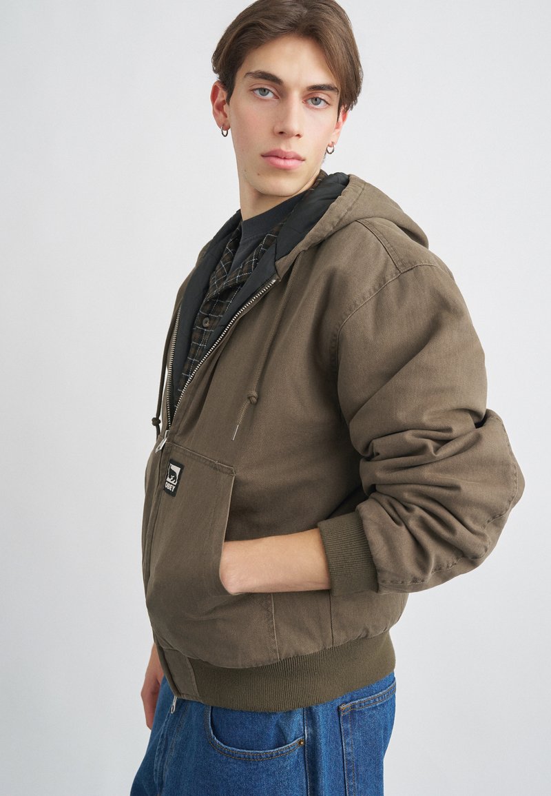 Smeđa jakna s kapuljačom i prednjim džepom, teksturiranog materijala i rebrastih manžeta. Nošena preko crne karirane košulje, s logotipom na prsima.