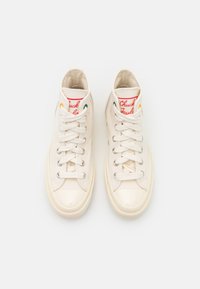 Pár krémově bílých vysokých plátěných tenisek s bílými tkaničkami, gumovými špičkami a červeným štítkem "Chuck Taylor" na jazycích.