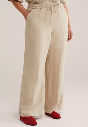 Beige Schlaghose mit elastischem Bund und Kordelzug, getragen mit roten flachen Schuhen, Hände des Models entspannt an den Seiten.