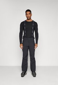 Maglietta nera a maniche lunghe abbinata a pantaloni scuri con bretelle; l'outfit è elegante, con una texture liscia e un design robusto. Stivali da sci neri completano il look.