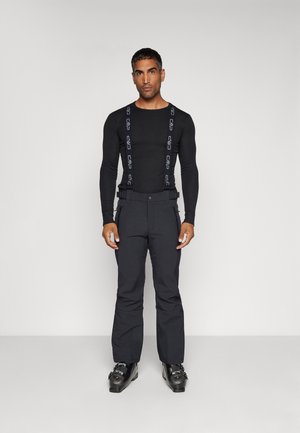 Maglietta nera a maniche lunghe abbinata a pantaloni scuri con bretelle; l'outfit è elegante, con una texture liscia e un design robusto. Stivali da sci neri completano il look.