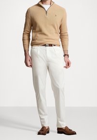 Homme portant un pull beige à fermeture quart de zip, un pantalon blanc plissé, une ceinture en cuir marron, des mocassins à glands marron et une montre marron au poignet gauche.