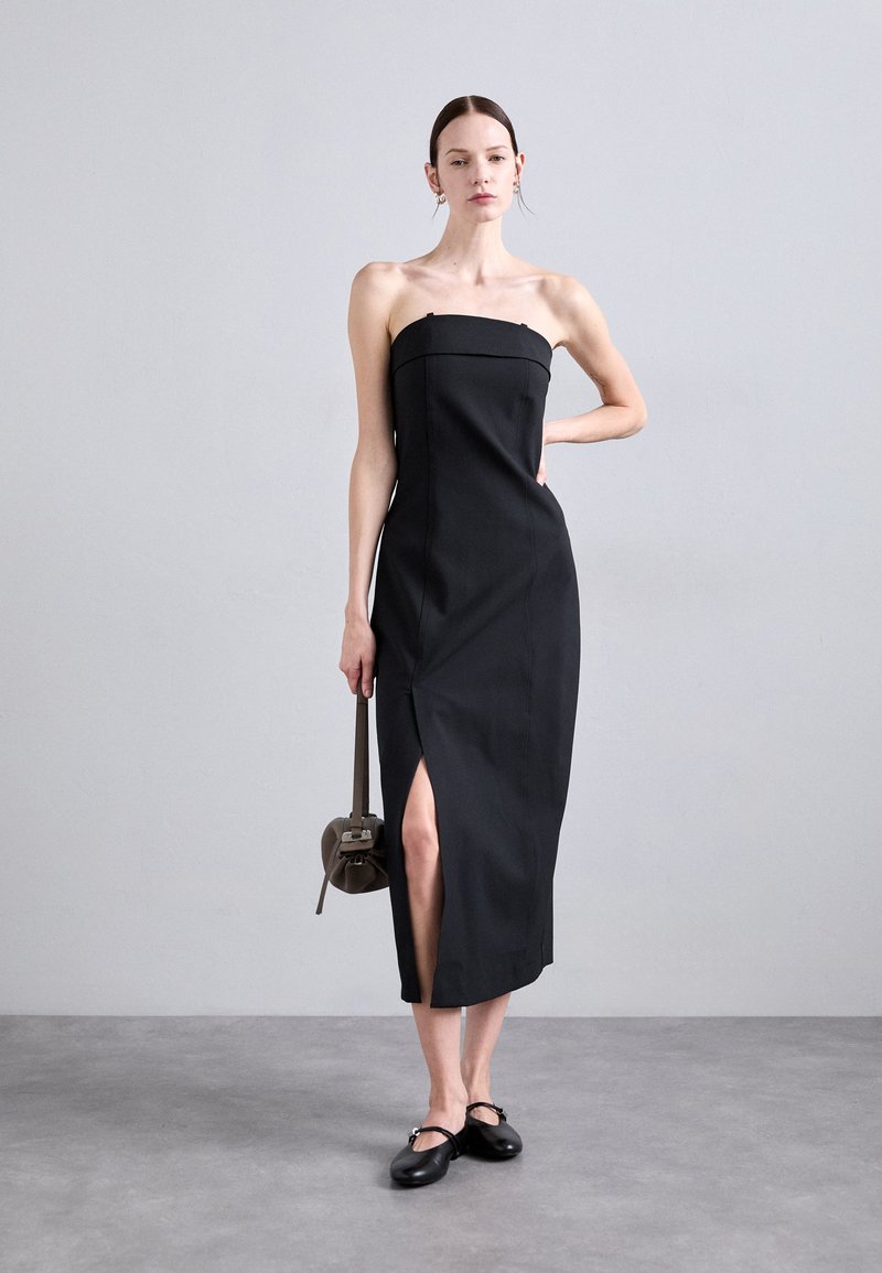 Black strapless dress with a fitted bodice, side slit, and knee-length hem. Model holds a brown shoulder bag, wearing black flats. Neutral background.