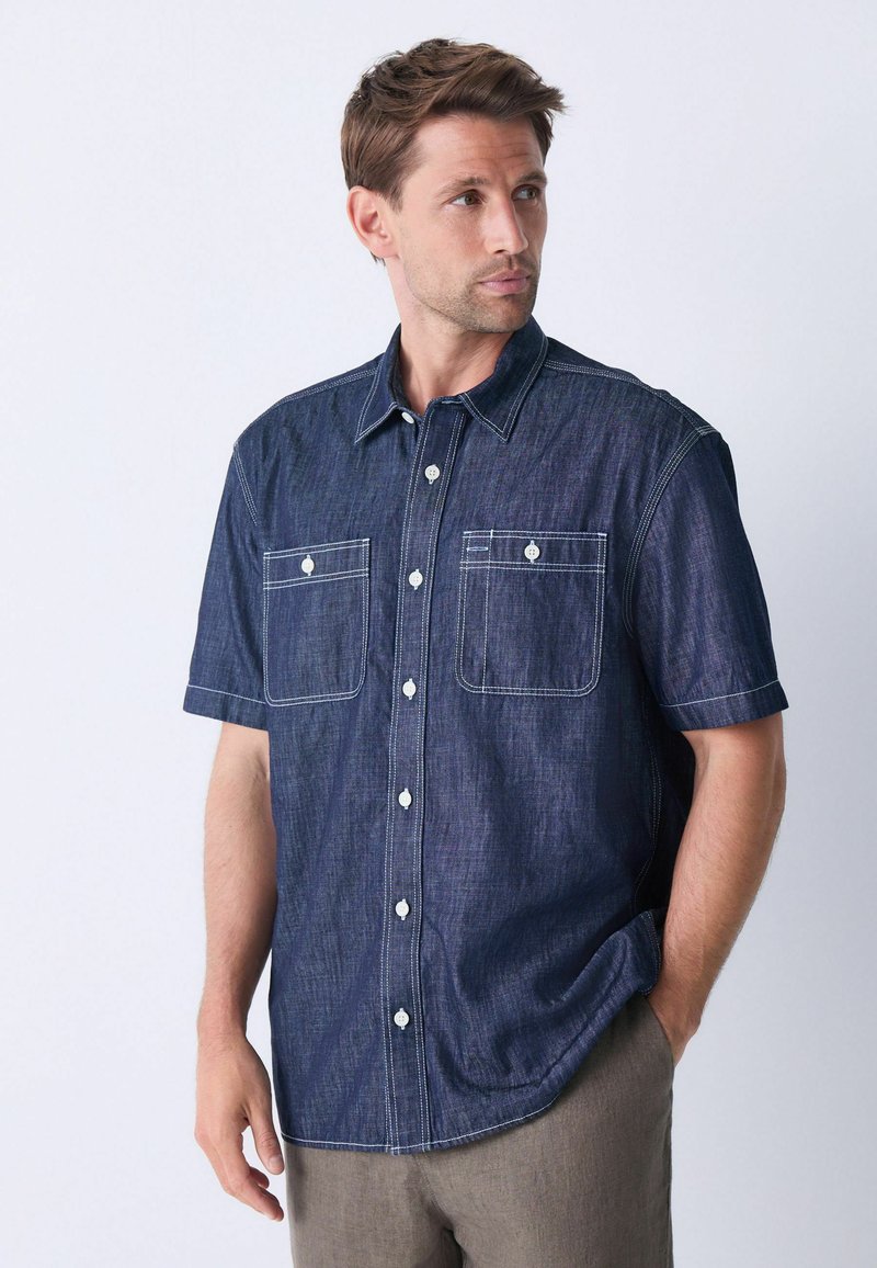 Camisa de denim de manga corta en azul oscuro con costuras blancas y dos bolsillos en el pecho, con cuello y cierre de botones.